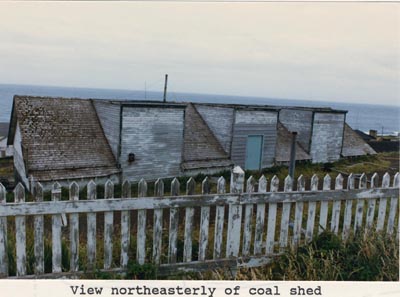 Photo of the northeasterly view of the coal shed, a long building with an alternating flat and angled roof and a picket fence.